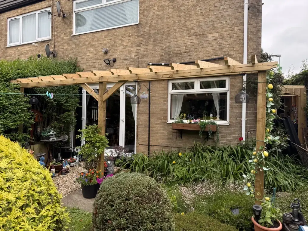 A timber pergola along the back of a house, over French doors and a planted garden