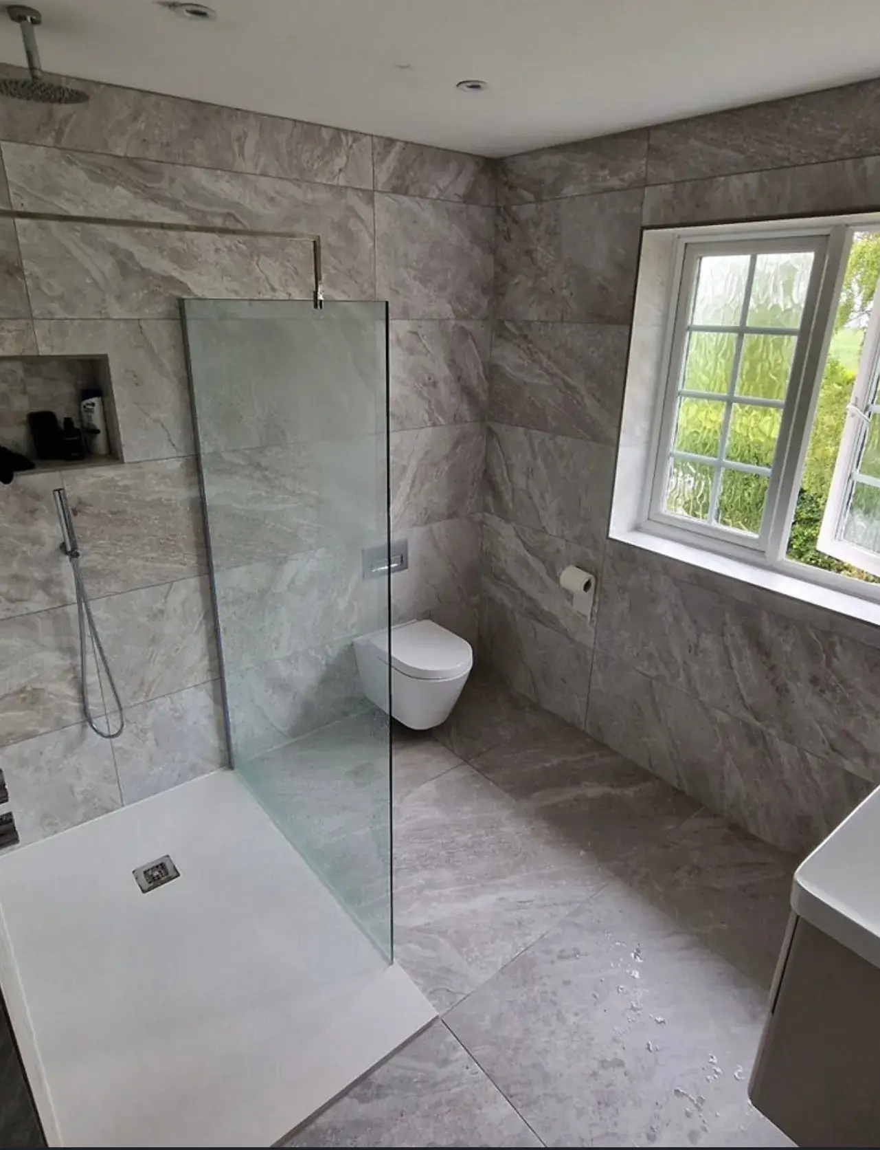 A walk-in shower with a glass screen and a wall-hung toilet, in grey stone-effect tiles