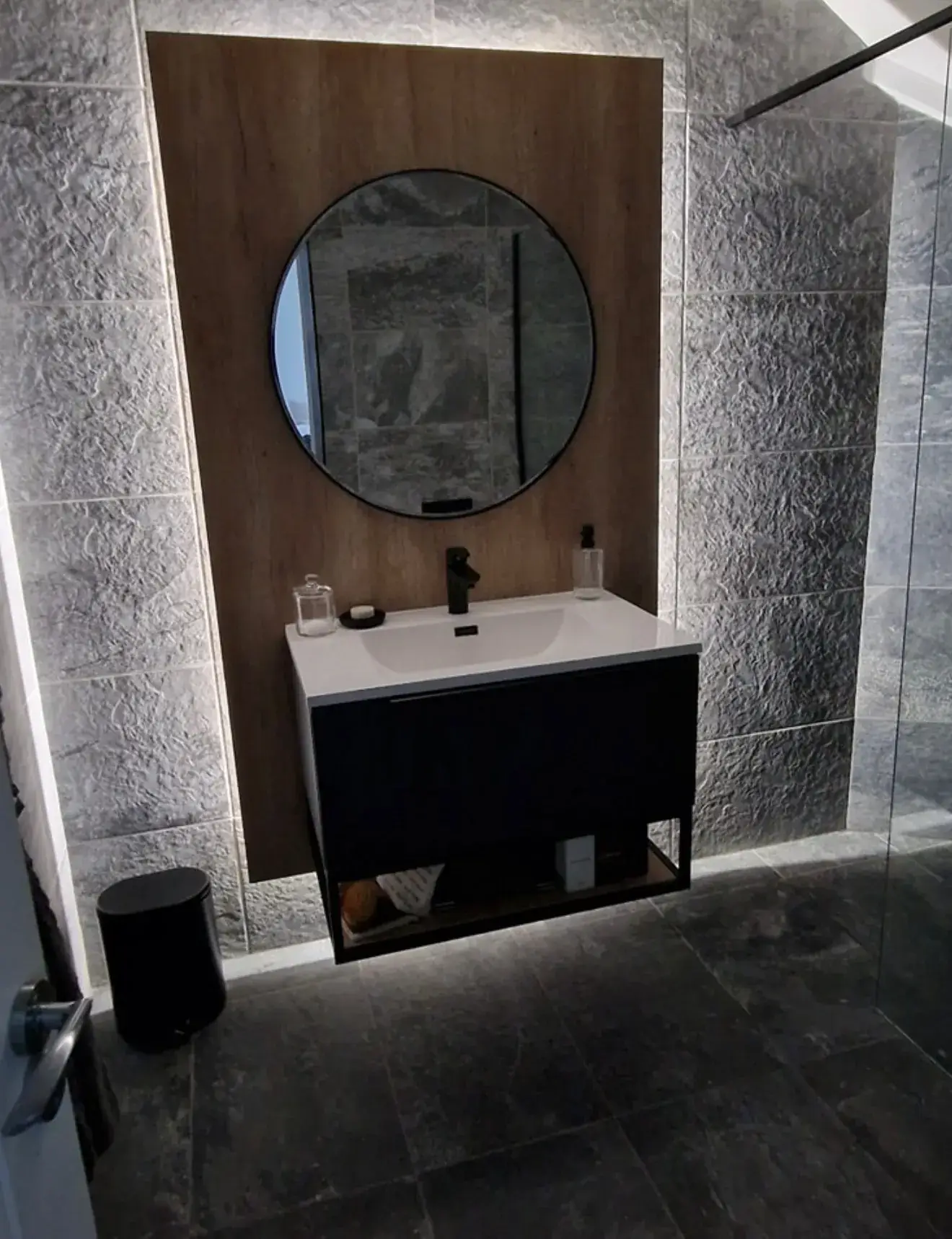 A wall-hung vanity unit with a round mirror on an oak-effect panel, in a grey tiled bathroom