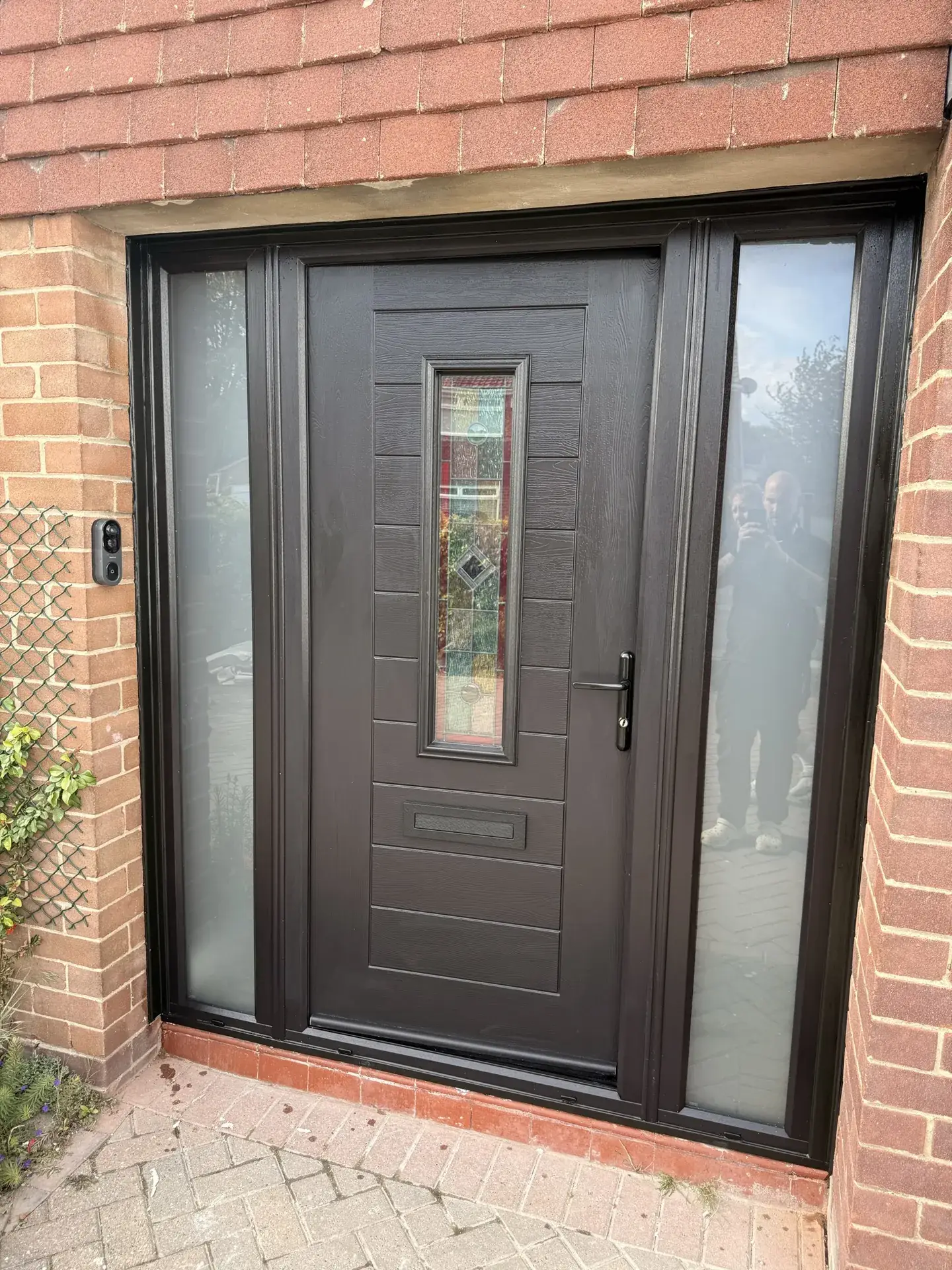 A dark grey composite front door with glazed side panels, in a red brick porch